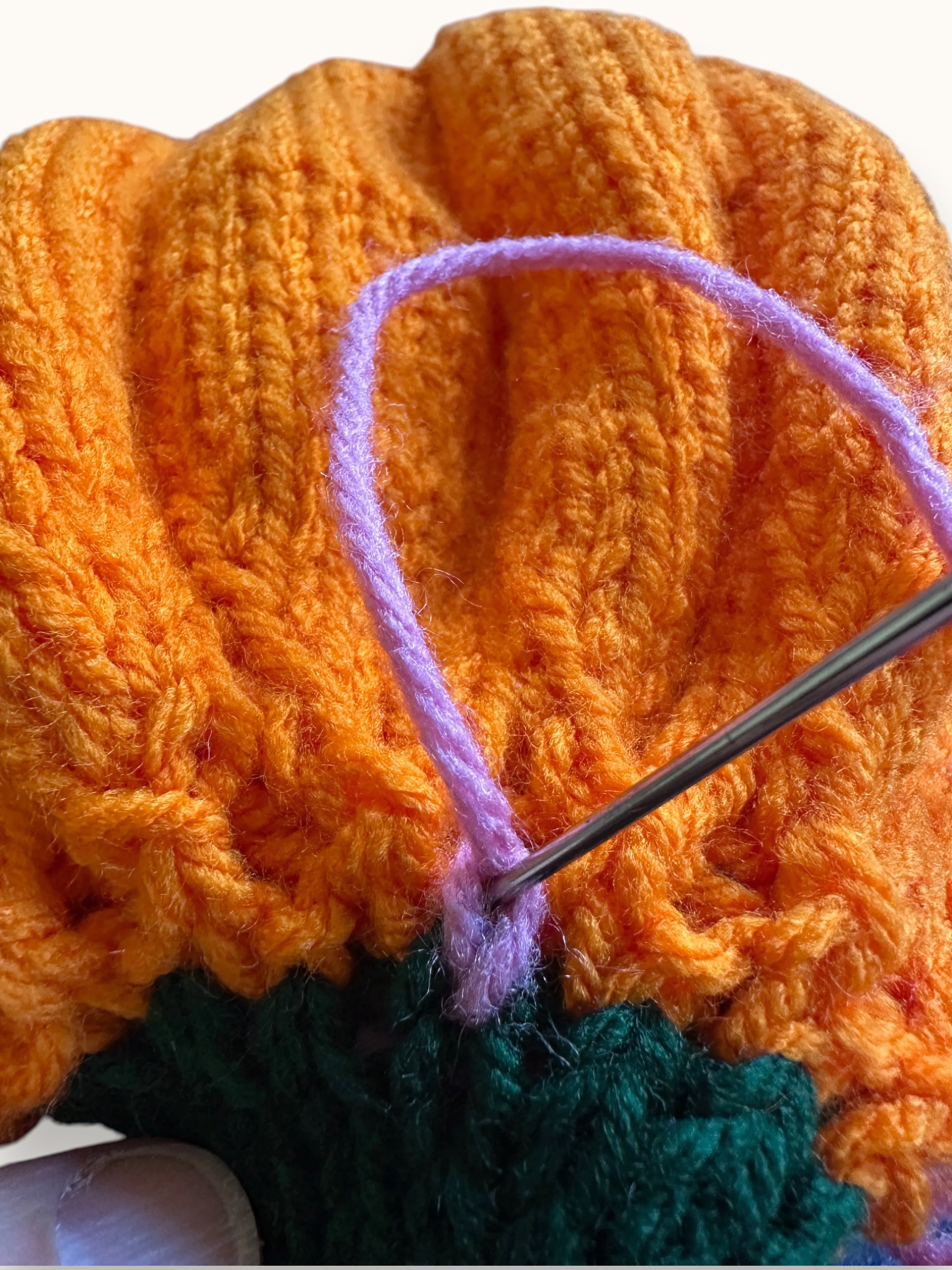 Close-up of a crocheted or knitted textile with a bright orange ribbed upper section, a dark green lower section, and a lavender loop being joined with a metal crochet hook.