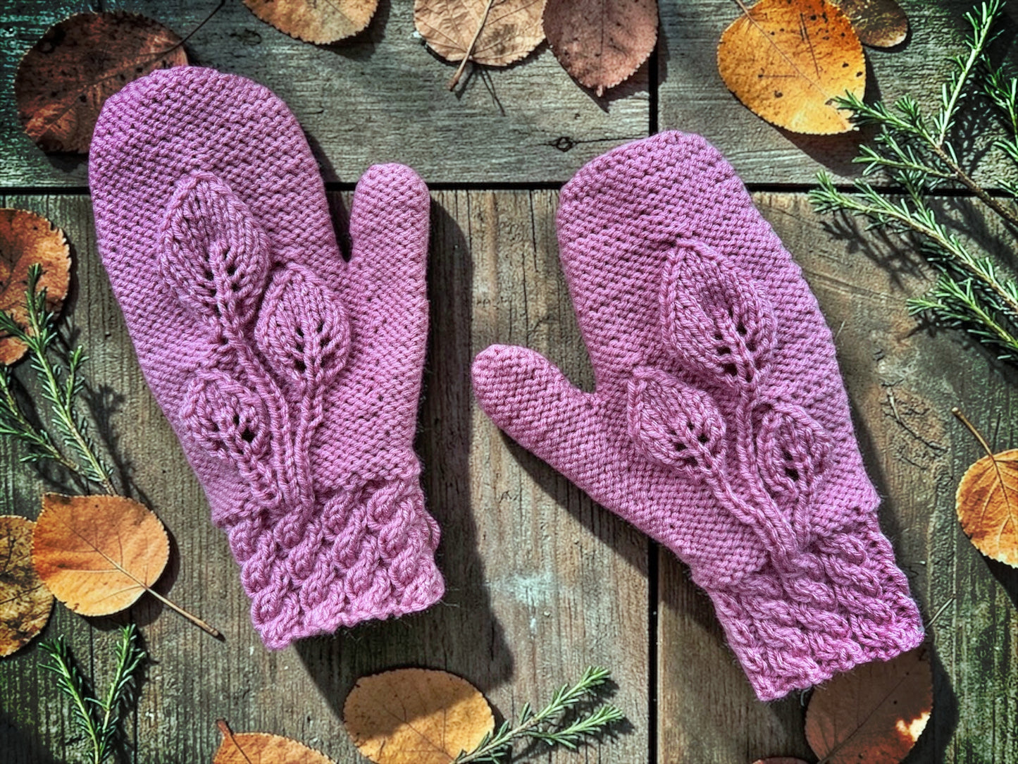 Pair of purple knitted mittens on a wooden surface with autumn leaves and branches.