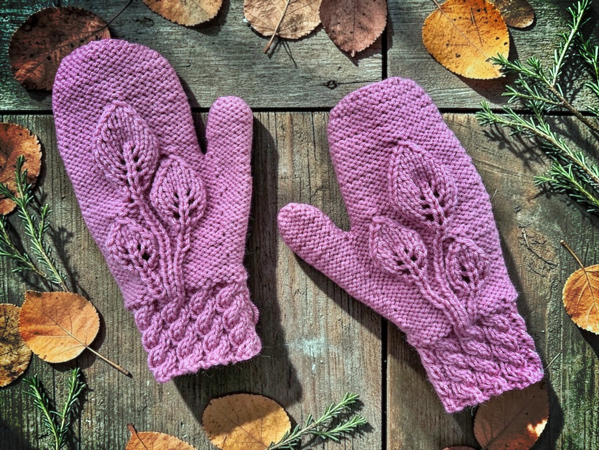 Pair of purple knitted mittens on a wooden surface with autumn leaves and branches.