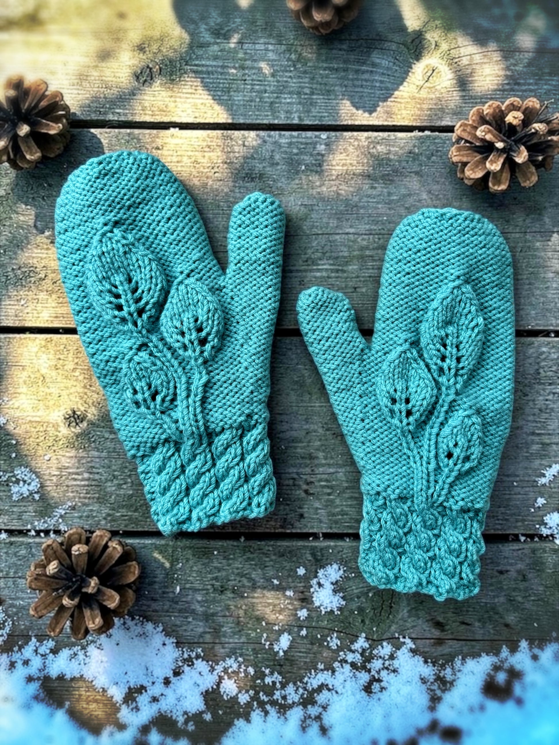 Pair of teal knitted mittens on a wooden surface with pine cones and snow. knitting pattern