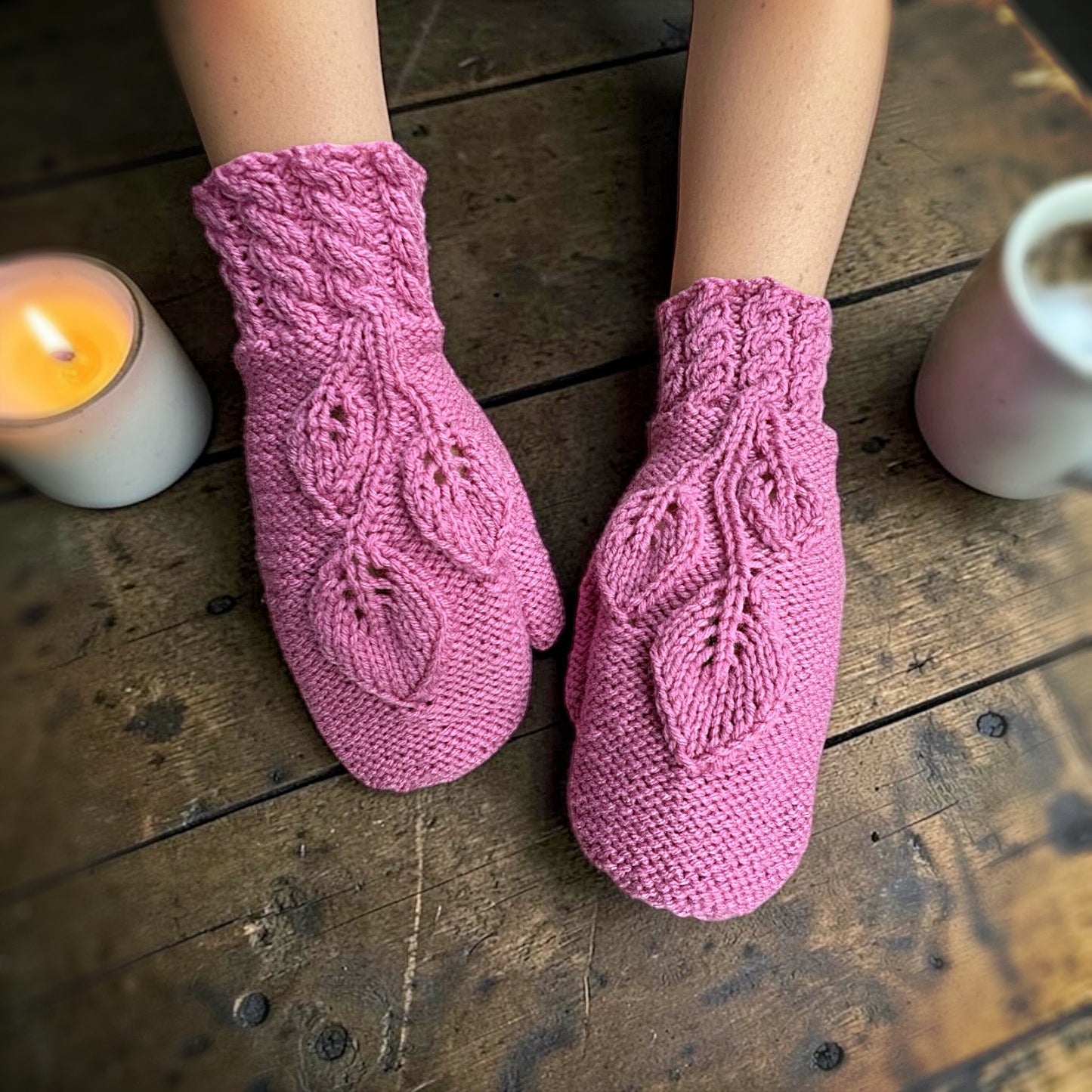 Pink knitted mittens on a wooden floor with a lit candle and mug in the background.