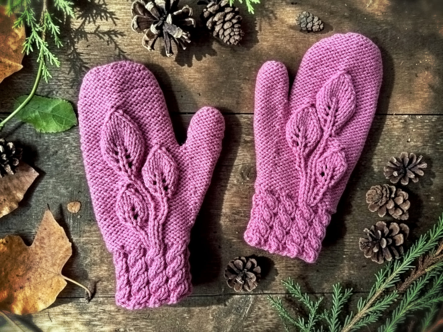 Pair of purple knitted mittens on a wooden surface with pine cones and leaves.