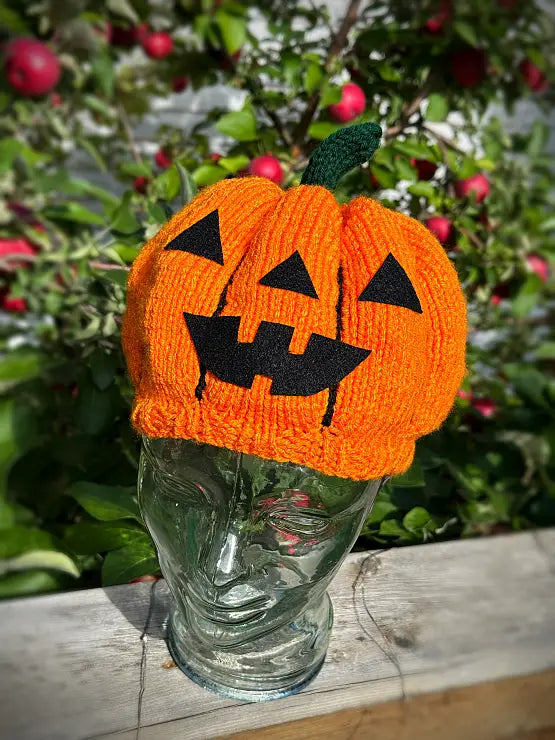 An orange knitted pumpkin hat with black embroidered jack-o'-lantern faces and a green stem, displayed on a clear glass face-shaped stand outdoors in front of leafy plants with red fruit.