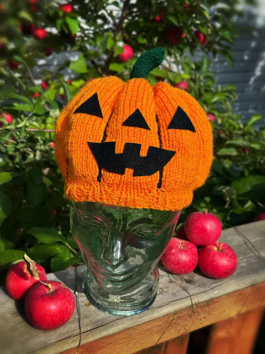 A bright orange knitted pumpkin hat with a black felt jack-o'-lantern face and a green stem, displayed on a clear glass mannequin head outdoors on a wooden surface with red apples and greenery in the background.
