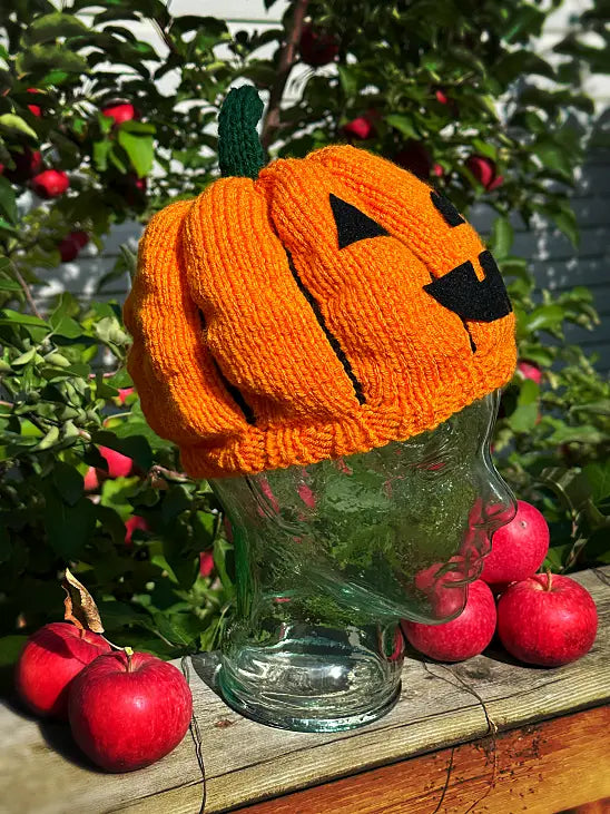 A bright orange knitted pumpkin hat with a green stem and black embroidered jack-o'-lantern face, displayed on a clear glass jar head outdoors on a wooden surface with red apples and leafy plants in the background.