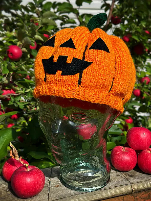 An orange knitted pumpkin hat with a black jack-o'-lantern face and green stem, displayed on a clear glass bottle head outdoors with red apples and greenery in the background.