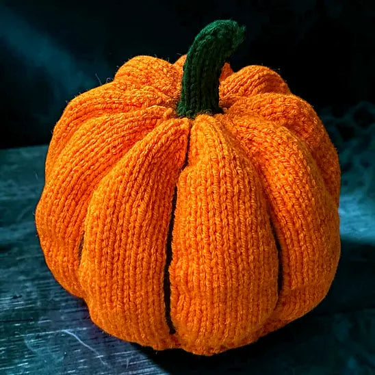 A knitted orange pumpkin decoration with a dark green stem, resting on a dark wooden surface with a smoky background.