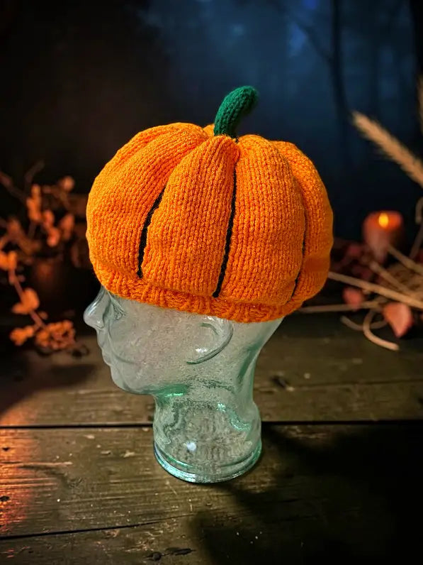 An orange knitted pumpkin hat with a green stem, displayed on a clear glass mannequin head against a dark, atmospheric background with autumn foliage.