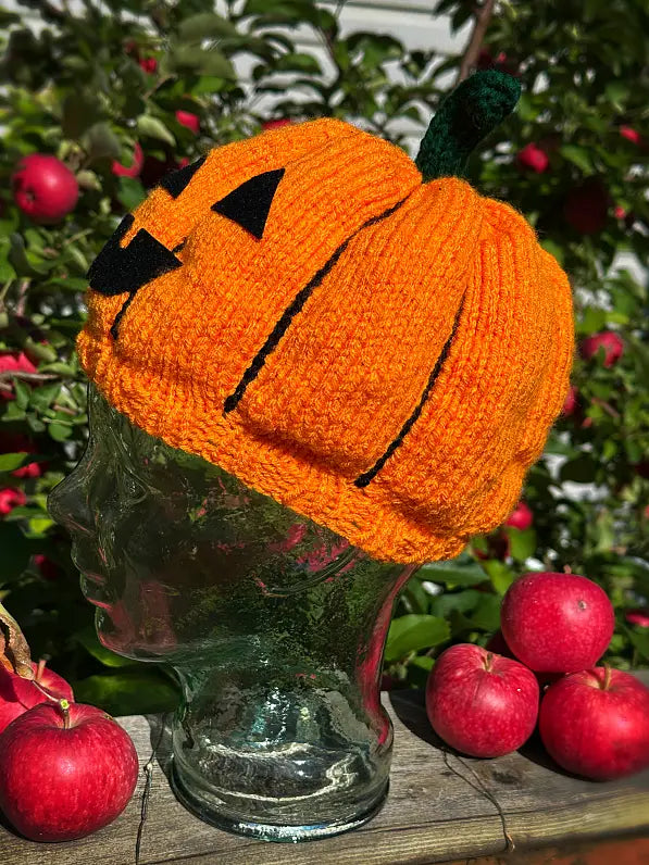 A bright orange knitted pumpkin hat with black stitched facial features and a dark green stem, displayed on a clear glass mannequin head outdoors among red apples and green foliage.