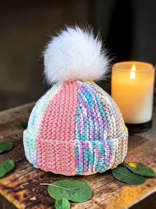 Knitted hat with a colorful pattern and white pom-pom on a wooden surface with a candle in the background.